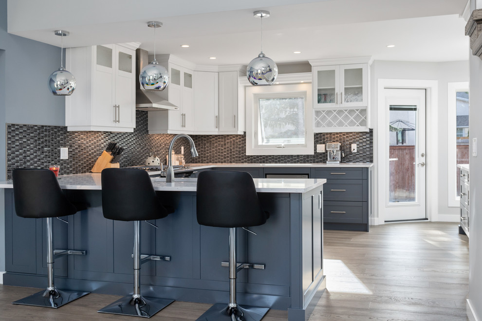 Bild på ett stort vintage vit vitt kök, med en undermonterad diskho, skåp i shakerstil, blå skåp, bänkskiva i kvarts, svart stänkskydd, stänkskydd i glaskakel, rostfria vitvaror, mellanmörkt trägolv, en köksö och grått golv