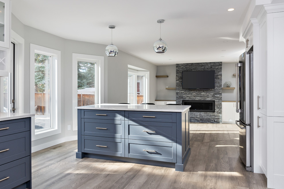 Klassisk inredning av ett stort vit vitt kök, med en undermonterad diskho, skåp i shakerstil, blå skåp, bänkskiva i kvarts, svart stänkskydd, stänkskydd i glaskakel, rostfria vitvaror, mellanmörkt trägolv, en köksö och grått golv