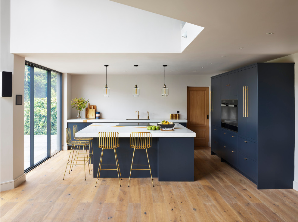 Moderne Küche in L-Form mit Kücheninsel, Unterbauwaschbecken, flächenbündigen Schrankfronten, blauen Schränken, Küchenrückwand in Weiß, schwarzen Elektrogeräten, braunem Holzboden, braunem Boden und weißer Arbeitsplatte