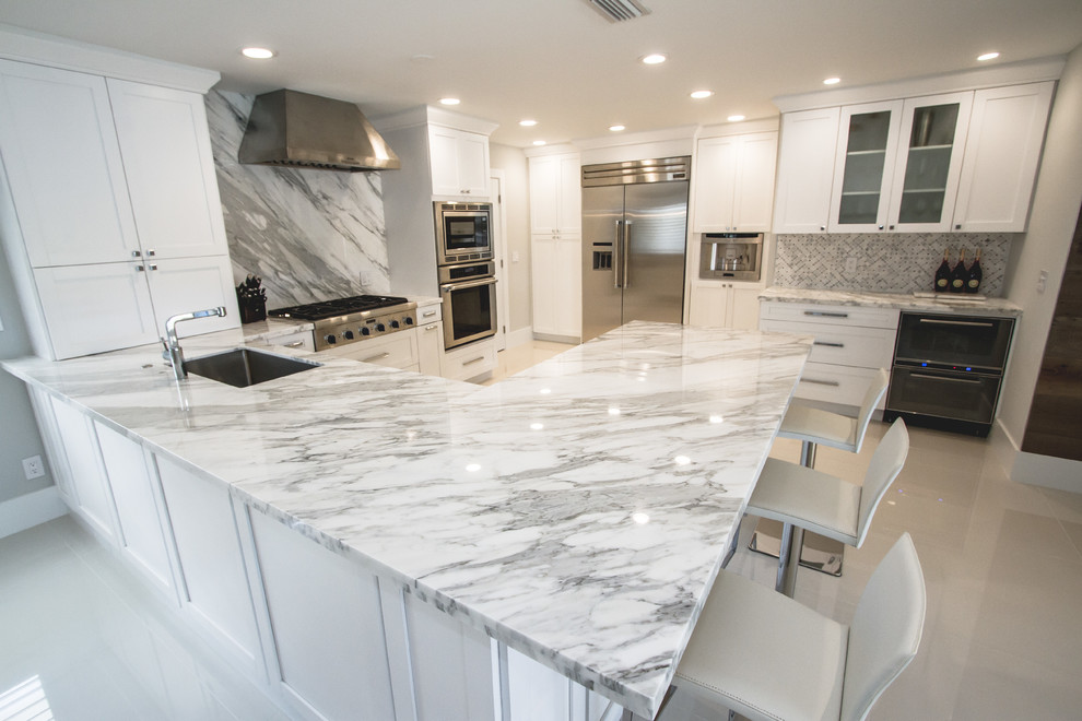 Modern inredning av ett avskilt, stort vit vitt l-kök, med en undermonterad diskho, skåp i shakerstil, vita skåp, marmorbänkskiva, rött stänkskydd, stänkskydd i marmor, rostfria vitvaror, klinkergolv i porslin, en halv köksö och beiget golv