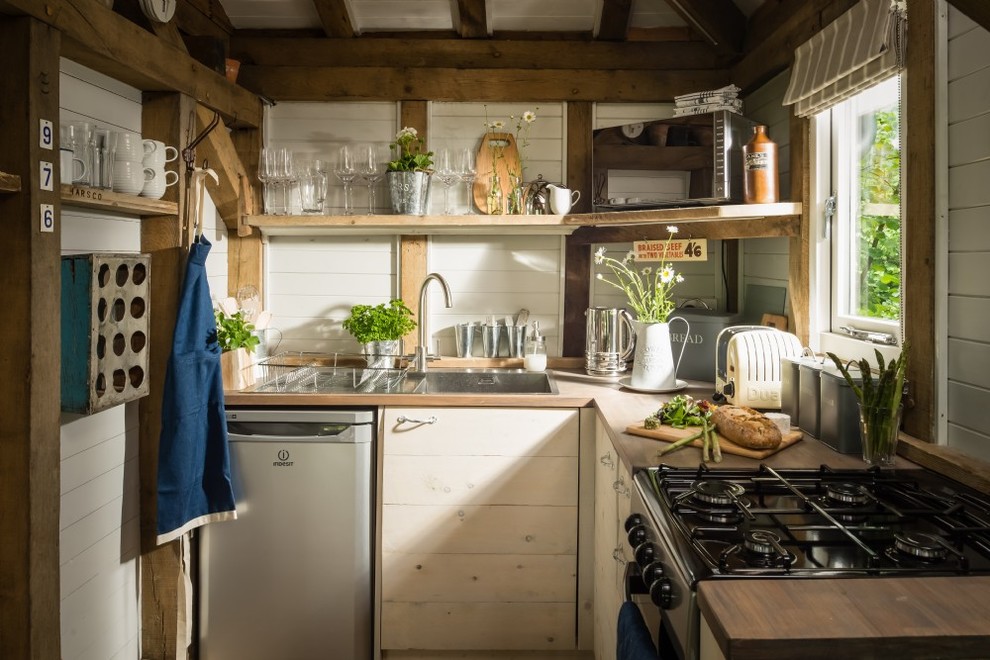 Small rustic l-shaped open plan kitchen in Sussex with a built-in sink, shaker cabinets, white cabinets, wood worktops, white splashback, wood splashback, stainless steel appliances, light hardwood flooring and beige floors.