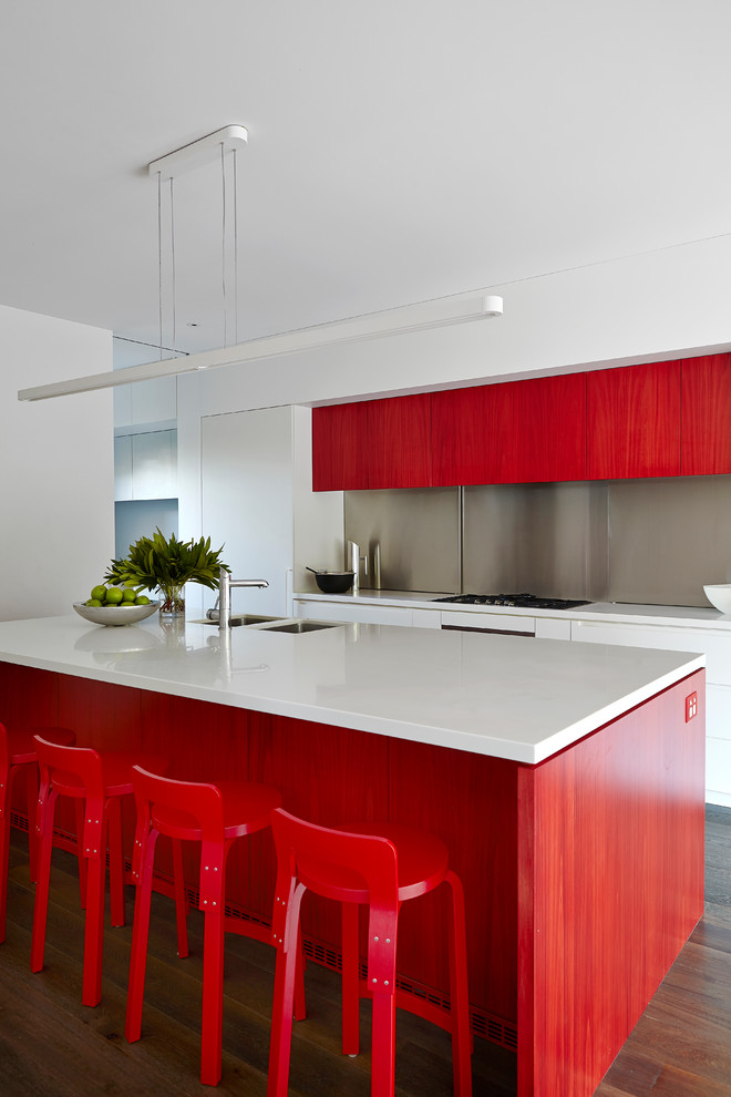 Idée de décoration pour une cuisine américaine parallèle design avec un évier encastré, un placard à porte plane, des portes de placard rouges, une crédence grise, un sol en bois brun, îlot, un sol marron et un plan de travail blanc.