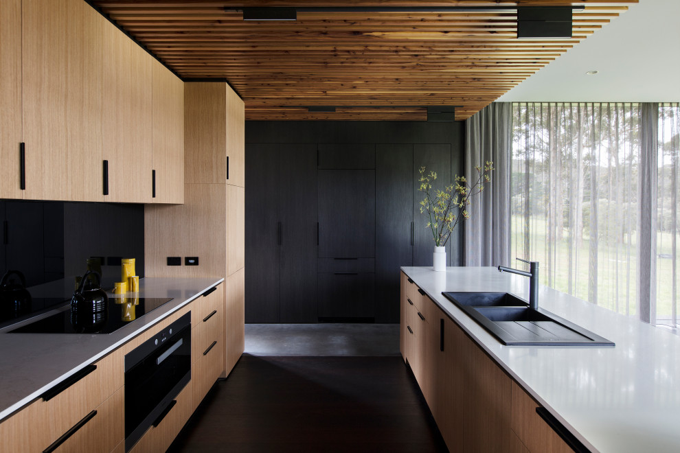 Moderne Küche in U-Form mit Doppelwaschbecken, hellen Holzschränken, schwarzen Elektrogeräten, dunklem Holzboden, flächenbündigen Schrankfronten, Kücheninsel und weißer Arbeitsplatte in Sonstige