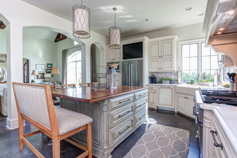 Inspiration for a large country grey and cream l-shaped open plan kitchen in Little Rock with raised-panel cabinets, an island, a belfast sink, composite countertops, grey splashback, stainless steel appliances, distressed cabinets, stone tiled splashback, concrete flooring and grey floors.