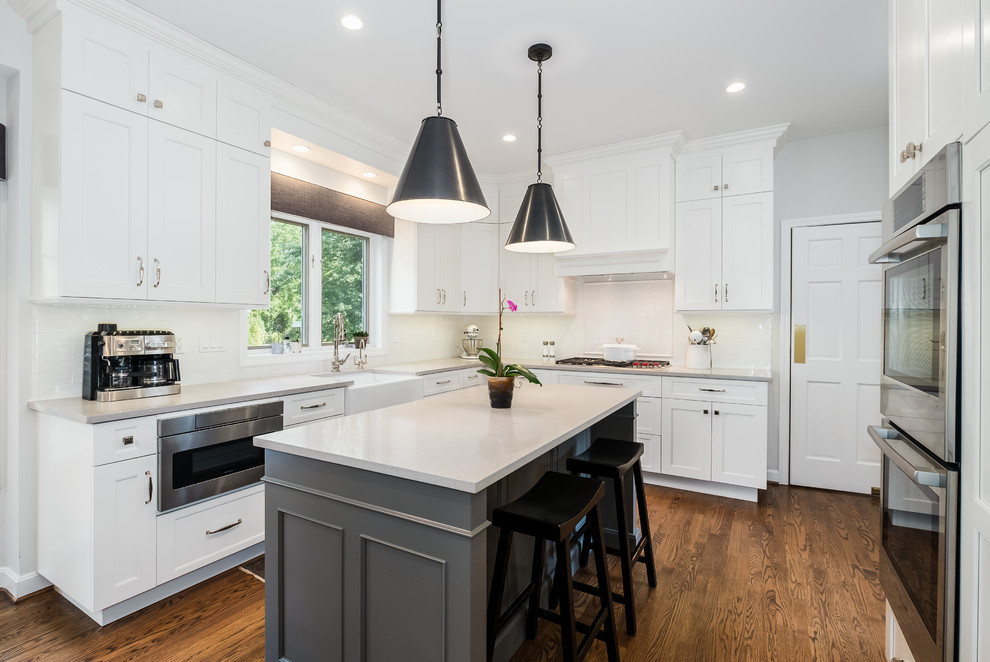 Mid-sized transitional l-shaped medium tone wood floor and brown floor eat-in kitchen photo in Bridgeport with a farmhouse sink, recessed-panel cabinets, white cabinets, quartz countertops, white backsplash, ceramic backsplash, stainless steel appliances and an island