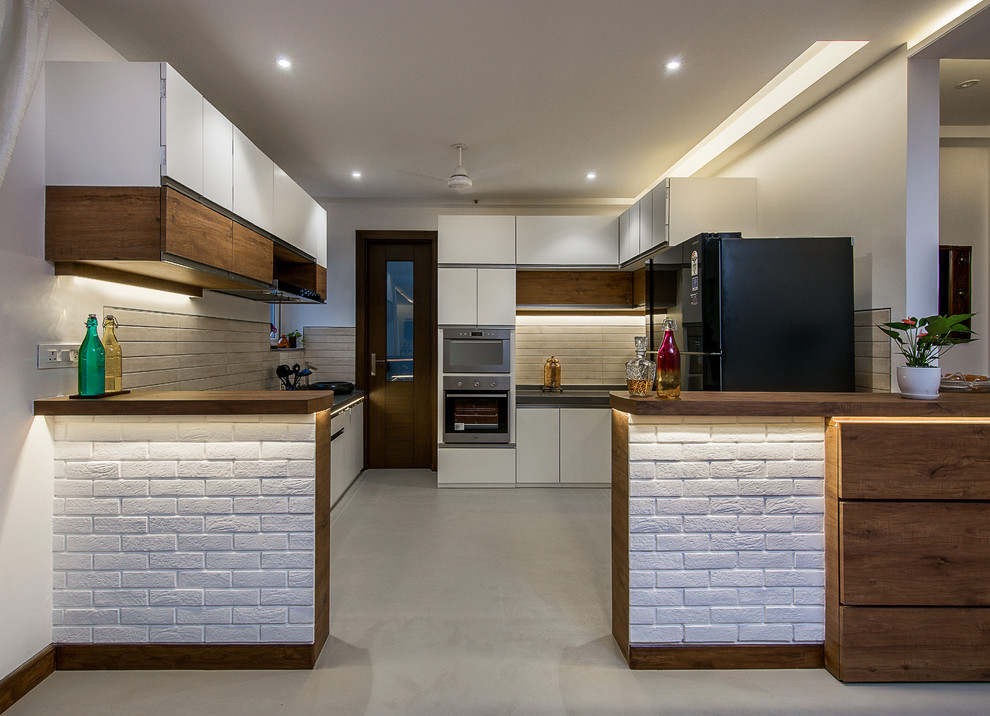 Inspiration for an eclectic gray floor kitchen remodel in Bengaluru with flat-panel cabinets, white cabinets, white backsplash, black appliances and a peninsula