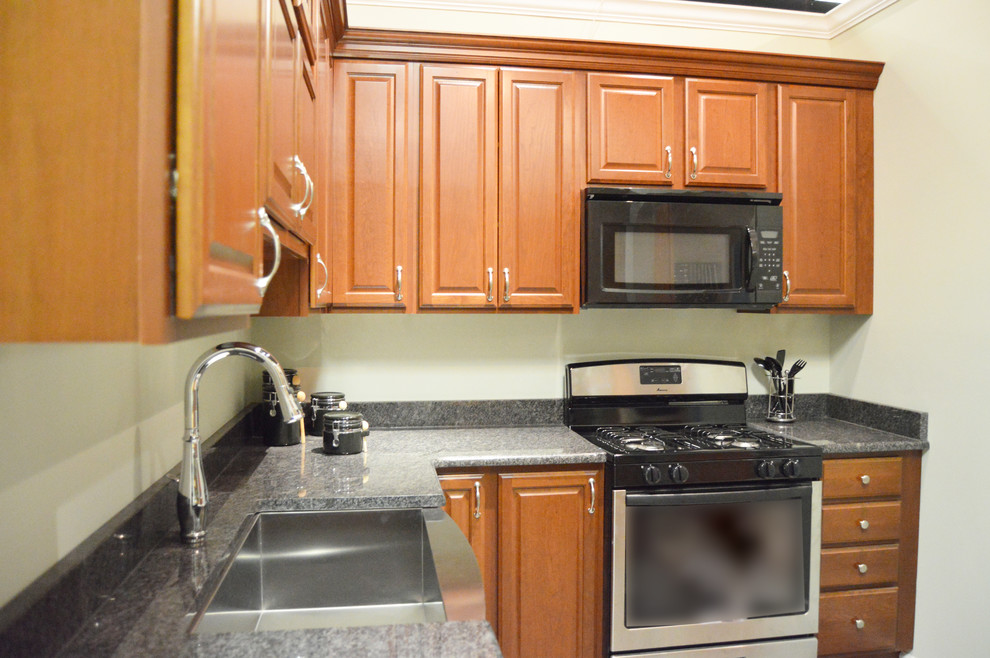 Cette photo montre une cuisine américaine chic en L et bois brun avec un évier de ferme, un placard avec porte à panneau surélevé, un plan de travail en granite et un électroménager noir.