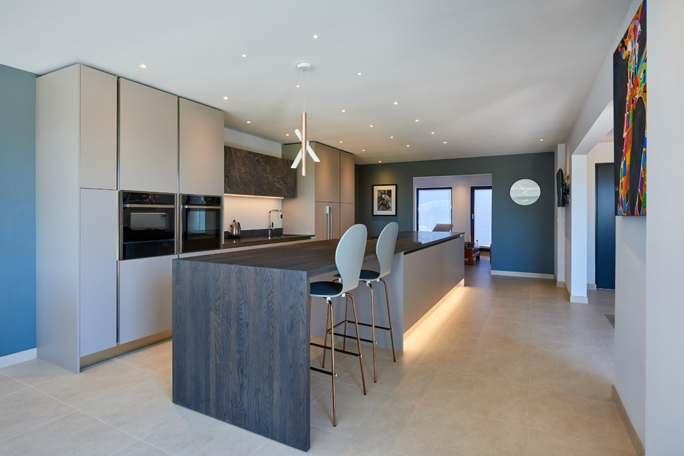 Large contemporary single-wall open plan kitchen in West Midlands with a built-in sink, flat-panel cabinets, grey cabinets, granite worktops, black appliances, limestone flooring, an island, beige floors and grey worktops.
