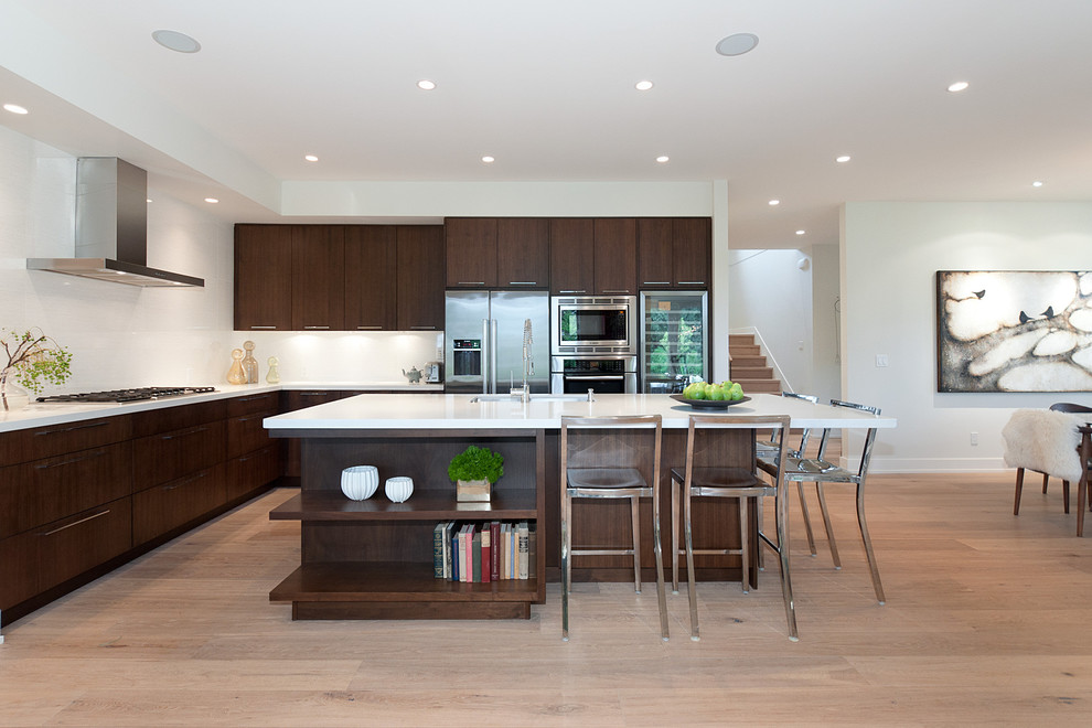 На фото: угловая кухня-гостиная в современном стиле с плоскими фасадами, темными деревянными фасадами, белым фартуком и техникой из нержавеющей стали