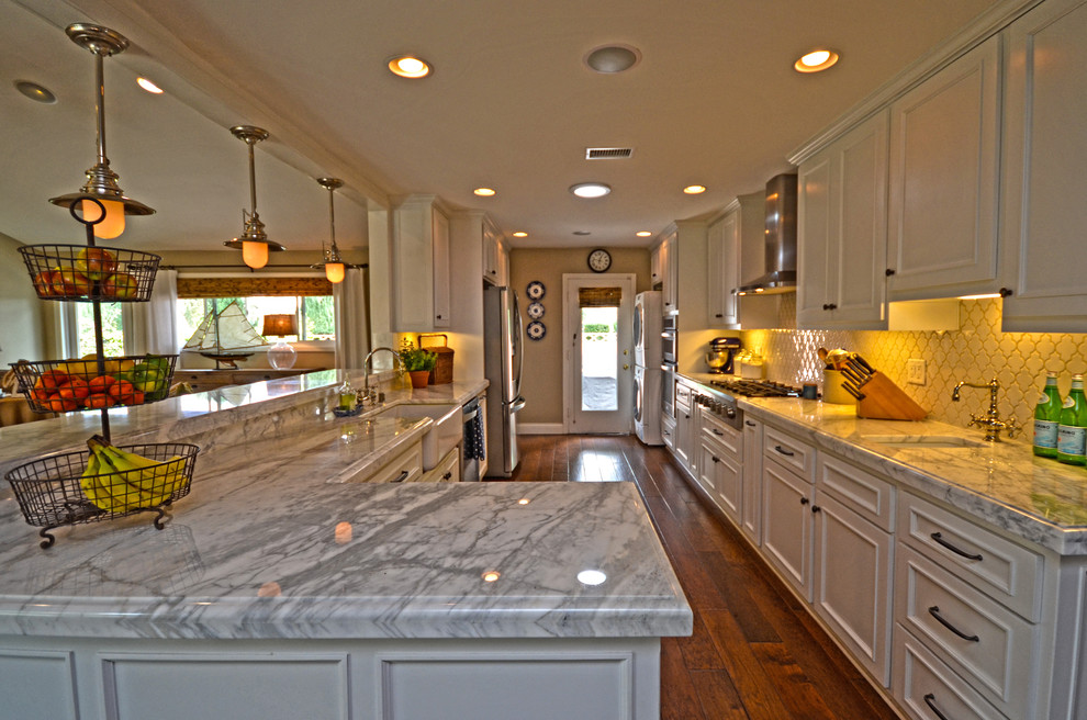Open concept kitchen - large traditional galley dark wood floor open concept kitchen idea in Orange County with a farmhouse sink, recessed-panel cabinets, white cabinets, marble countertops, white backsplash, ceramic backsplash, stainless steel appliances and a peninsula