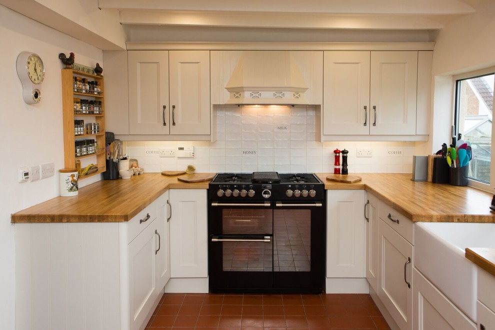 Bild på ett lantligt u-kök, med en rustik diskho, luckor med profilerade fronter, vita skåp, träbänkskiva, vitt stänkskydd, stänkskydd i keramik, svarta vitvaror och klinkergolv i terrakotta