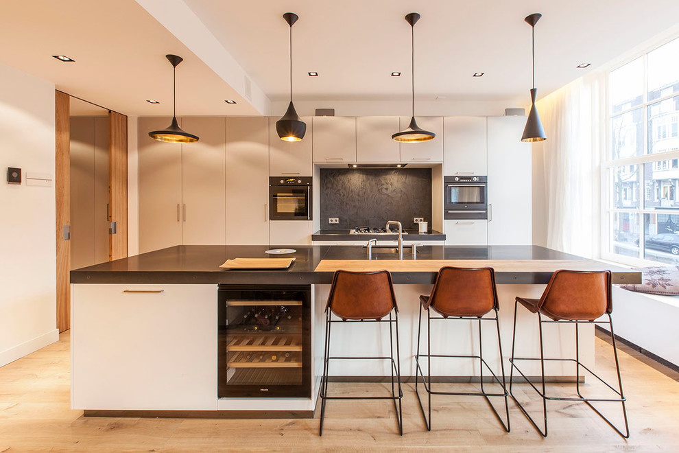 Bild på ett funkis kök, med en enkel diskho, släta luckor, beige skåp, svart stänkskydd, svarta vitvaror, ljust trägolv och en köksö