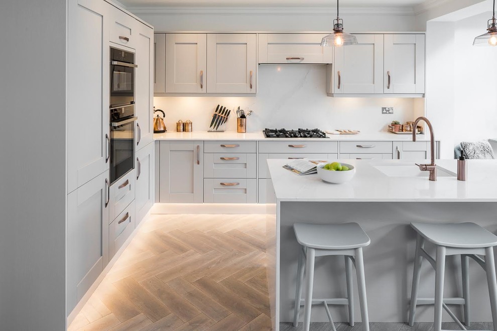 Mittelgroße Moderne Küche in L-Form mit Unterbauwaschbecken, Schrankfronten im Shaker-Stil, grauen Schränken, schwarzen Elektrogeräten, braunem Holzboden, Kücheninsel, braunem Boden und weißer Arbeitsplatte in London