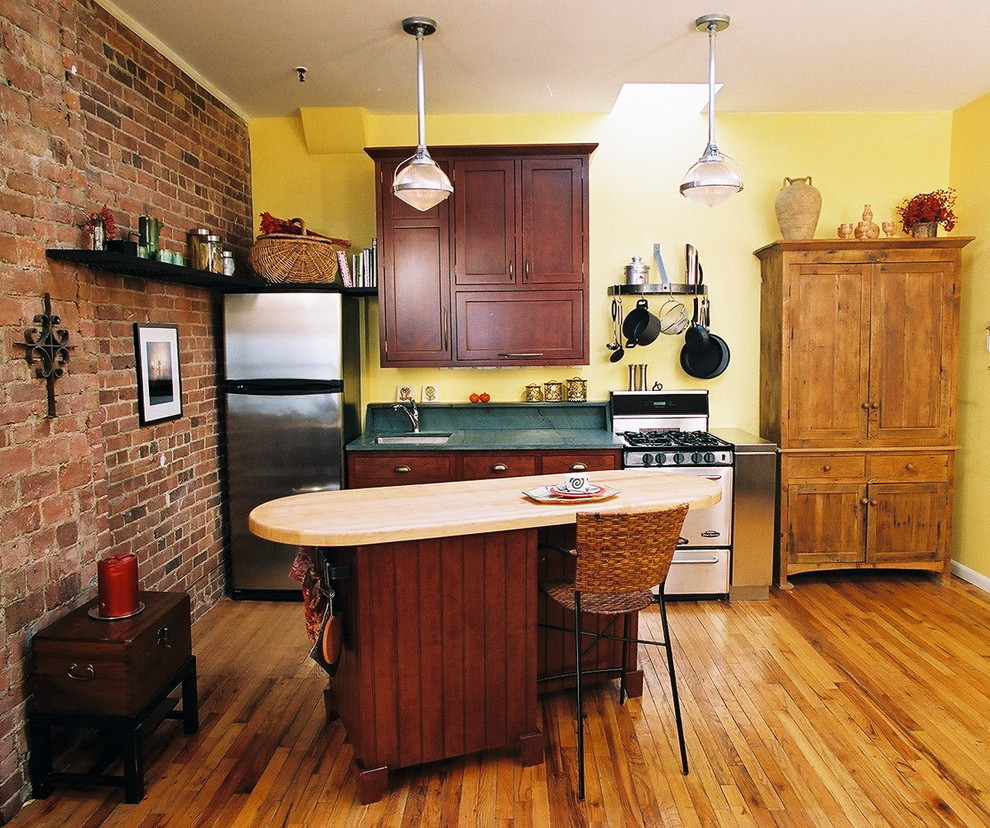 Cette photo montre une petite cuisine américaine linéaire éclectique en bois foncé avec un placard à porte affleurante, un plan de travail en bois, un électroménager en acier inoxydable, une crédence verte, un sol en bois brun et îlot.