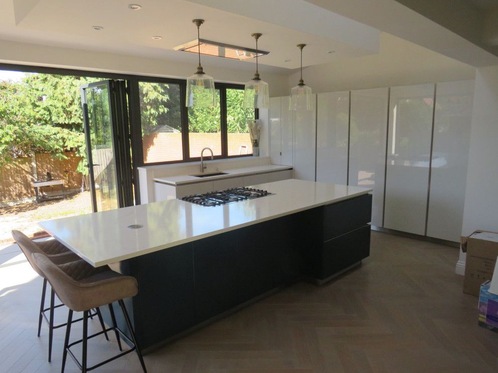 This is an example of a large contemporary l-shaped open plan kitchen in Essex with an integrated sink, flat-panel cabinets, blue cabinets, quartz worktops, white splashback, stainless steel appliances, light hardwood flooring, an island, beige floors and white worktops.