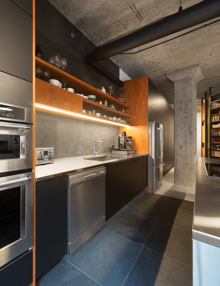 Design ideas for a medium sized industrial galley kitchen/diner in Montreal with a built-in sink, flat-panel cabinets, black cabinets, stainless steel worktops, grey splashback, slate splashback, stainless steel appliances, slate flooring, an island, grey floors and grey worktops.