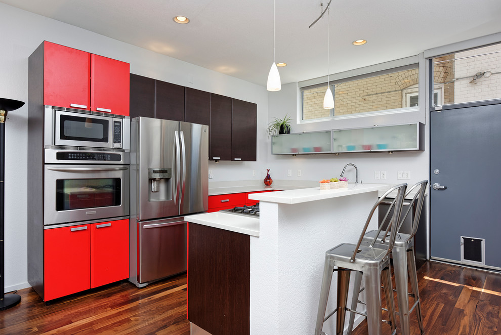 Exemple d'une cuisine tendance en L avec un placard à porte plane, des portes de placard rouges, une crédence blanche, un électroménager en acier inoxydable, un sol en bois brun, îlot, un sol marron et un plan de travail blanc.