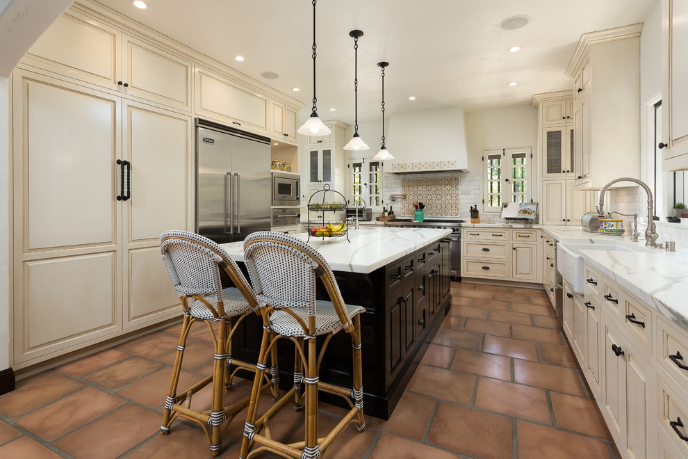 Inspiration for a mediterranean u-shaped terra-cotta tile kitchen remodel in Los Angeles with a farmhouse sink, raised-panel cabinets, beige cabinets, multicolored backsplash, stainless steel appliances, an island and marble countertops