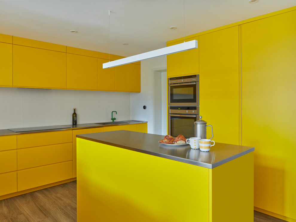 Moderne Küche mit Waschbecken, flächenbündigen Schrankfronten, gelben Schränken, Edelstahl-Arbeitsplatte, Küchengeräten aus Edelstahl, hellem Holzboden, Kücheninsel und beigem Boden in London
