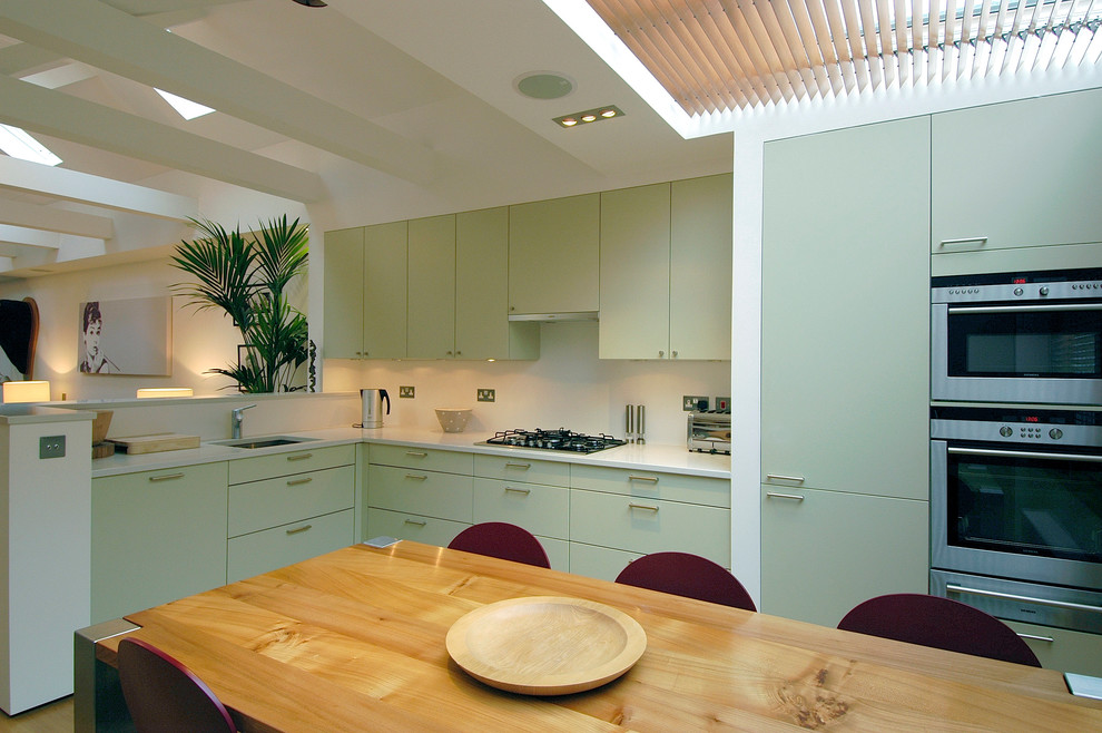 This is an example of a small classic u-shaped kitchen/diner in Hertfordshire with a built-in sink, flat-panel cabinets, green cabinets, composite countertops, white splashback, stainless steel appliances, light hardwood flooring and no island.
