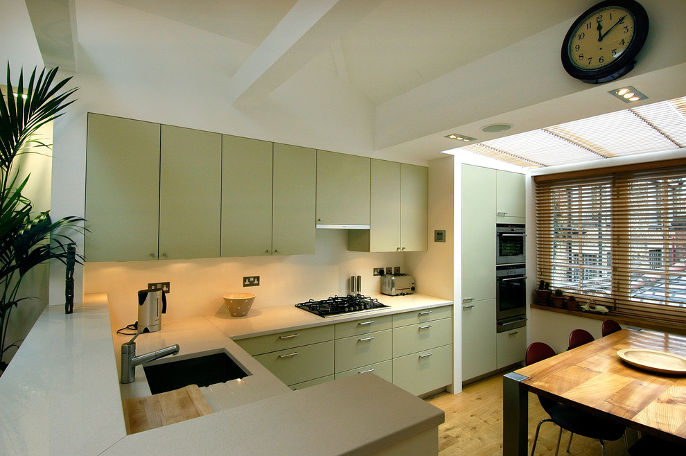 Photo of a small traditional u-shaped kitchen/diner in Hertfordshire with a built-in sink, flat-panel cabinets, green cabinets, composite countertops, white splashback, stainless steel appliances, light hardwood flooring and no island.