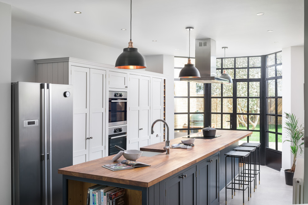 Classic galley kitchen in London with a belfast sink, shaker cabinets, grey cabinets, wood worktops, stainless steel appliances, an island, grey floors and brown worktops.