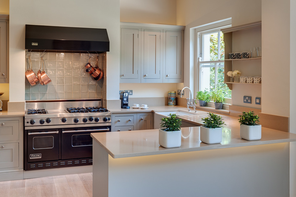 Example of a classic u-shaped light wood floor kitchen design in London with a farmhouse sink, shaker cabinets, gray cabinets, black appliances, a peninsula and gray backsplash