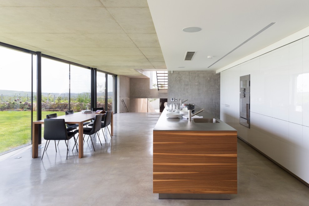 Идея дизайна: большая параллельная кухня в стиле модернизм с обеденным столом, врезной мойкой, плоскими фасадами, белыми фасадами, бетонным полом, островом и техникой из нержавеющей стали