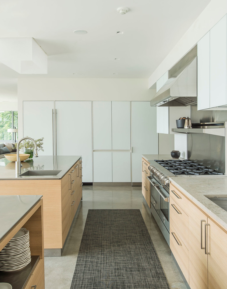 Modern inredning av ett kök, med en enkel diskho, släta luckor, skåp i ljust trä, stänkskydd med metallisk yta, rostfria vitvaror, betonggolv och flera köksöar