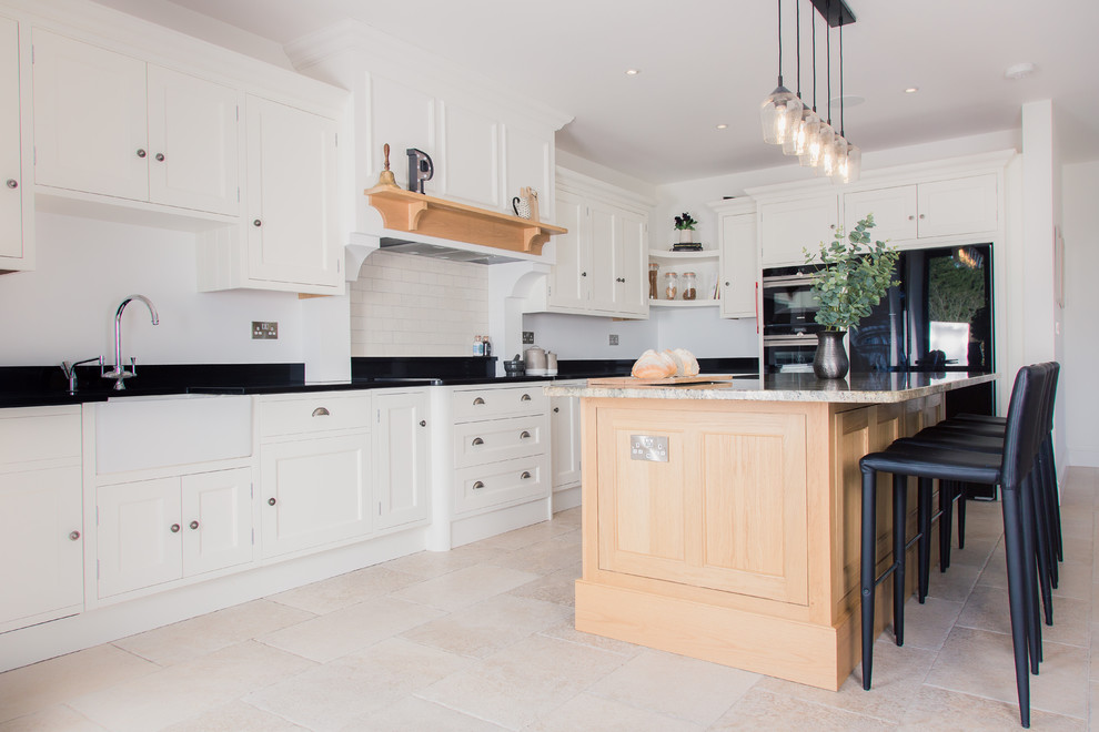 Example of a transitional l-shaped beige floor open concept kitchen design in Other with a farmhouse sink, shaker cabinets, white cabinets, white backsplash, black appliances, an island and black countertops