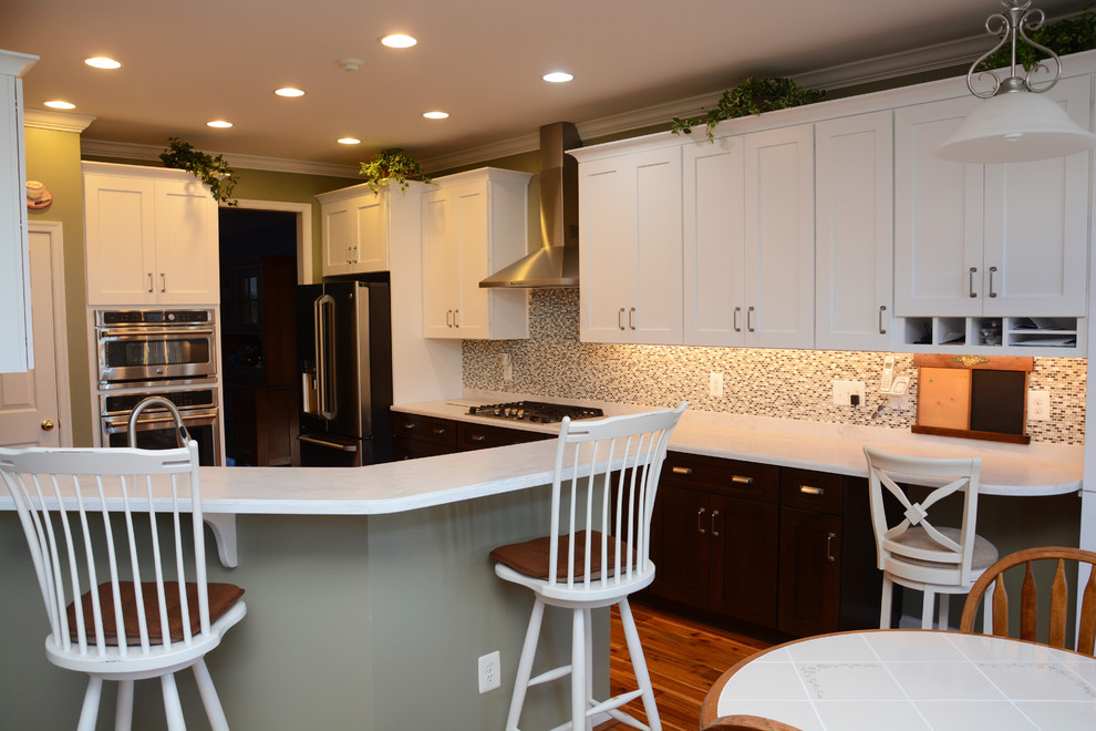 Photo of a large traditional u-shaped open plan kitchen in DC Metro with a belfast sink, shaker cabinets, white cabinets, composite countertops, multi-coloured splashback, mosaic tiled splashback, stainless steel appliances, medium hardwood flooring, no island, brown floors and white worktops.