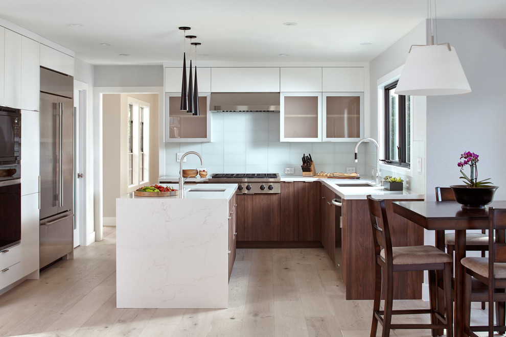 Photo of a medium sized modern l-shaped kitchen/diner in San Francisco with a built-in sink, glass-front cabinets, dark wood cabinets, engineered stone countertops, green splashback, glass tiled splashback, stainless steel appliances, light hardwood flooring, an island, grey floors and white worktops.