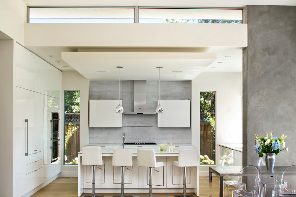 Cette image montre une cuisine encastrable design en L avec un placard à porte plane, des portes de placard blanches, une crédence grise, un sol en bois brun et îlot.