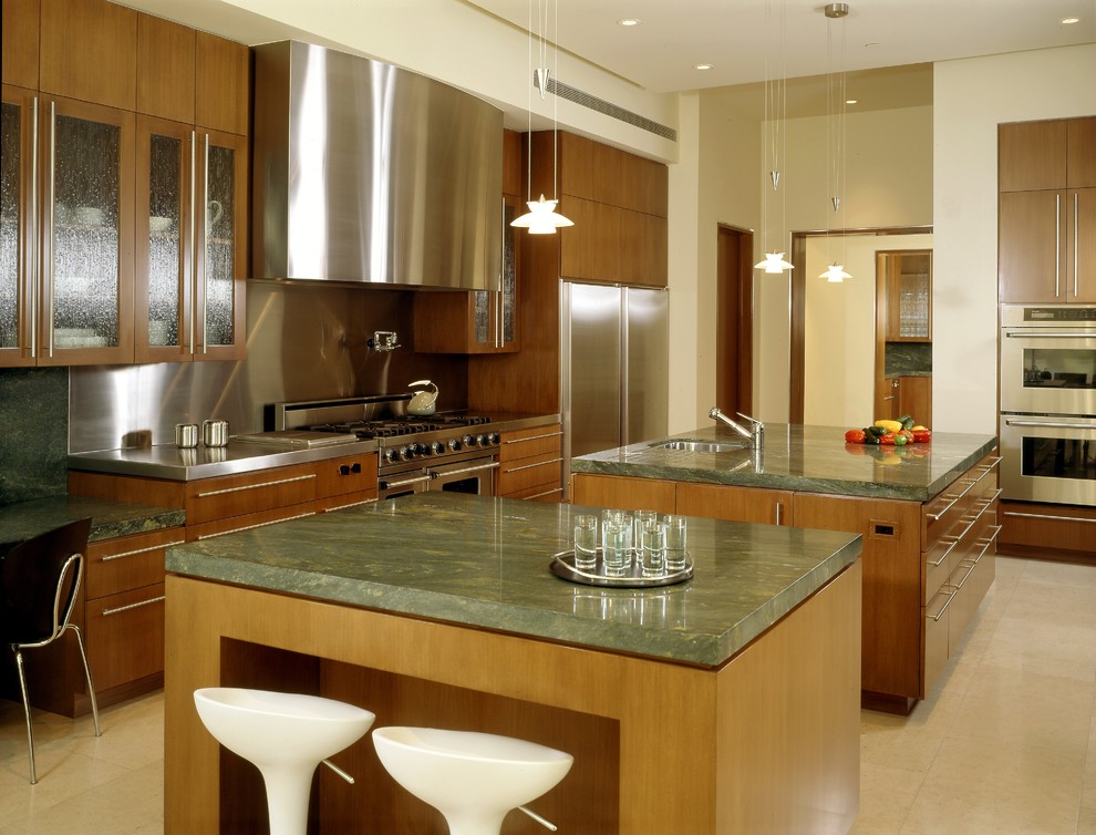 Cette image montre une cuisine design en bois brun avec un placard à porte vitrée, un électroménager en acier inoxydable, un sol en calcaire, un évier encastré, une crédence métallisée et 2 îlots.