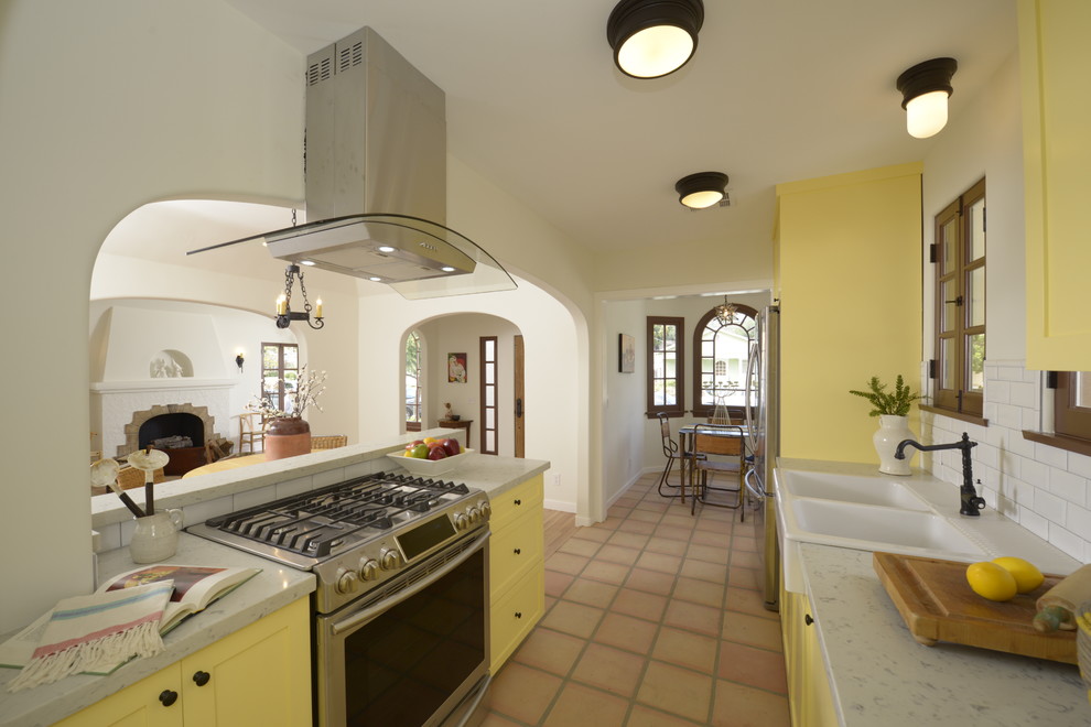 Foto på ett mellanstort medelhavsstil kök, med en halv köksö, skåp i shakerstil, gula skåp, bänkskiva i kvarts, vitt stänkskydd, stänkskydd i tunnelbanekakel, integrerade vitvaror, en rustik diskho och klinkergolv i terrakotta