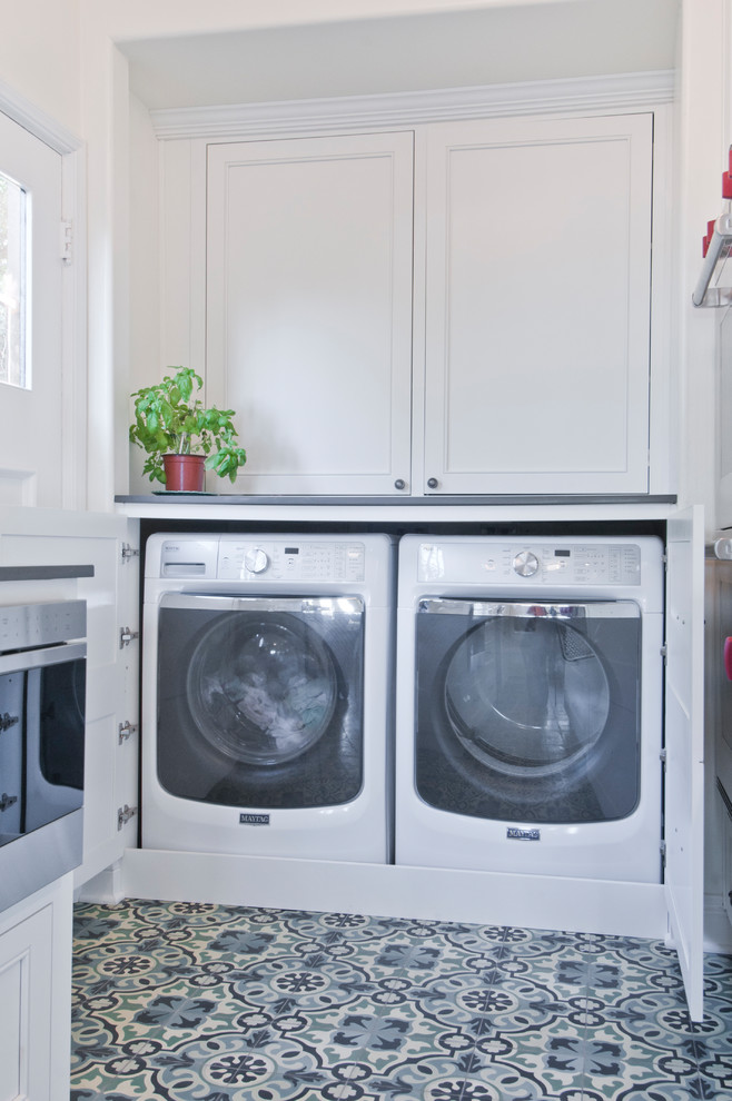 Klassisk inredning av en mellanstor grå l-formad grått tvättstuga, med en undermonterad diskho, luckor med infälld panel, vita skåp, bänkskiva i kvarts, grönt stänkskydd, stänkskydd i keramik, klinkergolv i porslin och flerfärgat golv