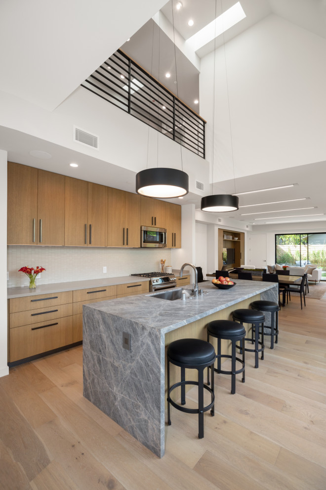 Example of a trendy galley light wood floor and beige floor kitchen design in Los Angeles with a single-bowl sink, flat-panel cabinets, light wood cabinets, quartzite countertops, white backsplash, porcelain backsplash, stainless steel appliances, an island and gray countertops