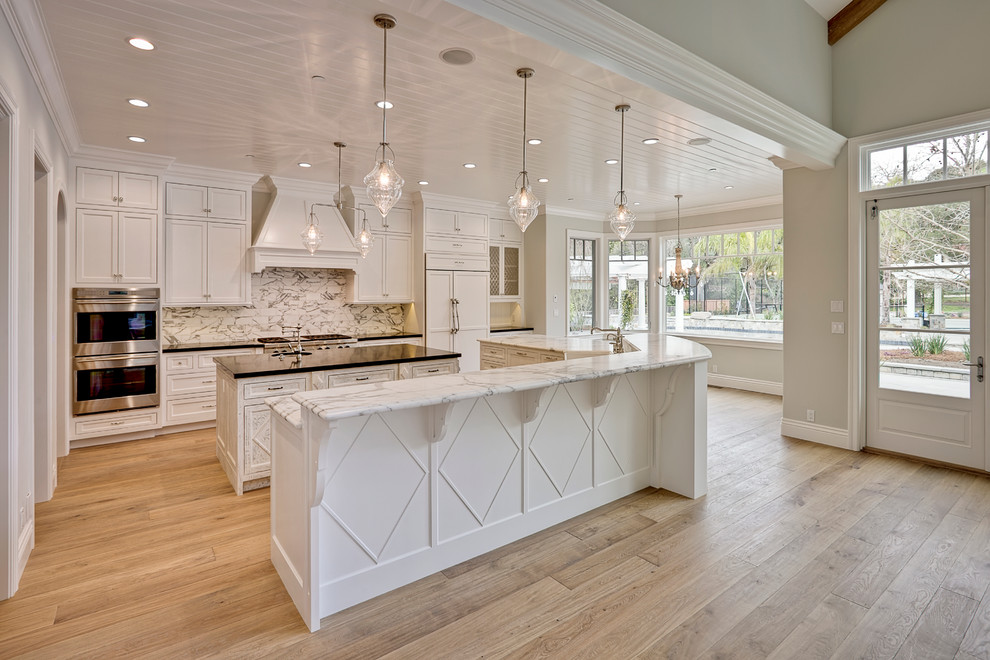 Large elegant u-shaped light wood floor kitchen photo in San Francisco with a farmhouse sink, recessed-panel cabinets, white cabinets, marble countertops, white backsplash, stone slab backsplash, stainless steel appliances and two islands