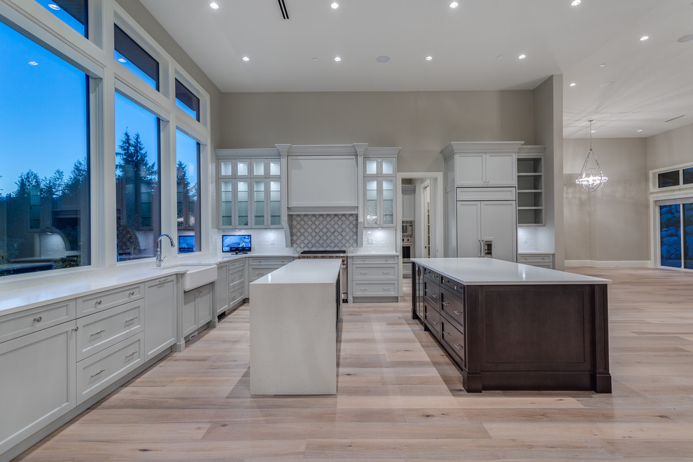 This is an example of an expansive modern l-shaped open plan kitchen in Vancouver with a submerged sink, shaker cabinets, white cabinets, engineered stone countertops, white splashback, travertine splashback, white appliances, medium hardwood flooring, multiple islands and beige floors.