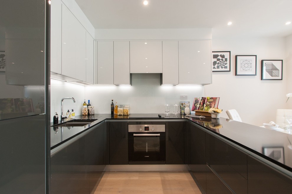 ロンドンにある小さなコンテンポラリースタイルのおしゃれなキッチン (ダブルシンク、フラットパネル扉のキャビネット、黒いキャビネット、ガラス板のキッチンパネル、シルバーの調理設備、淡色無垢フローリング、アイランドなし、ベージュの床) の写真