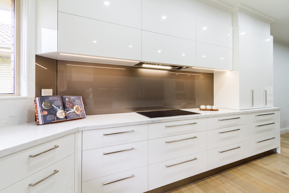 Large trendy u-shaped light wood floor and brown floor eat-in kitchen photo in Melbourne with a drop-in sink, flat-panel cabinets, white cabinets, quartz countertops, metallic backsplash, glass sheet backsplash, black appliances, an island and white countertops