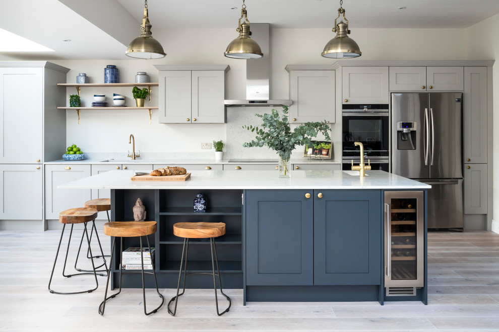 This is an example of a large traditional kitchen in London with shaker cabinets, quartz worktops, an island, a submerged sink, grey cabinets, stainless steel appliances, light hardwood flooring and white worktops.