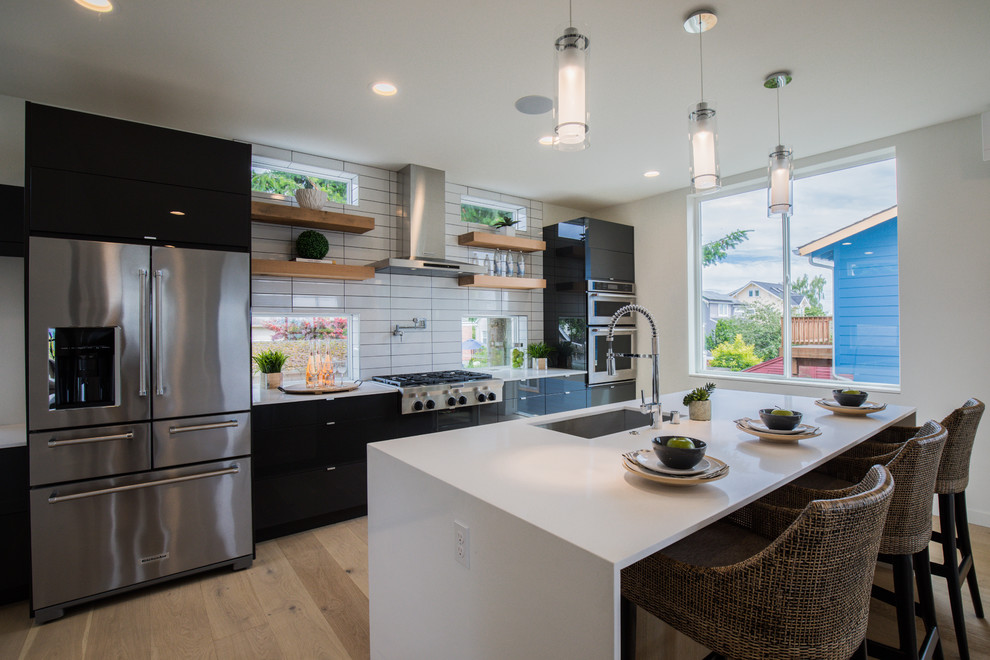Example of a trendy galley medium tone wood floor and brown floor kitchen design in Seattle with an undermount sink, flat-panel cabinets, black cabinets, white backsplash, stainless steel appliances, an island and white countertops