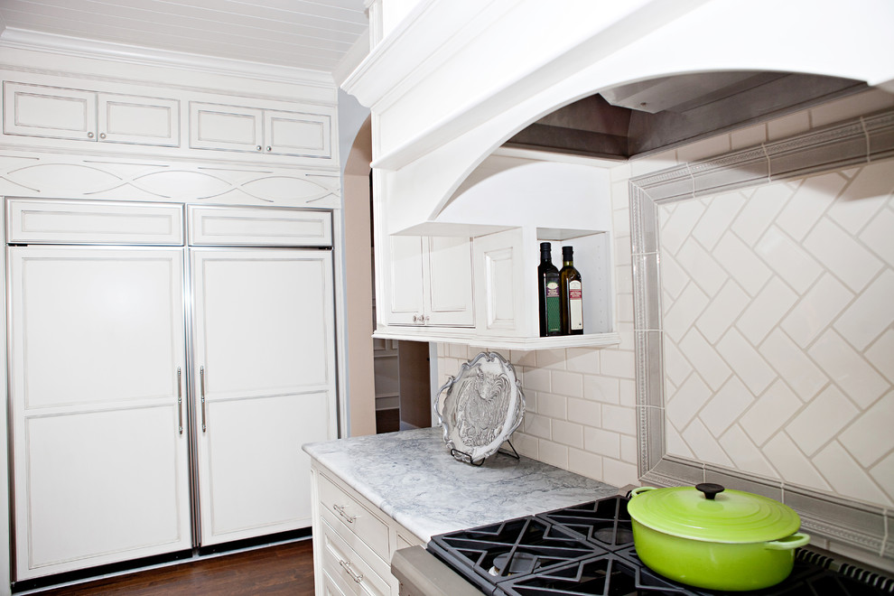 Foto på ett stort vintage grå kök, med en undermonterad diskho, luckor med infälld panel, vita skåp, marmorbänkskiva, vitt stänkskydd, stänkskydd i tunnelbanekakel, integrerade vitvaror, mörkt trägolv, en köksö och brunt golv