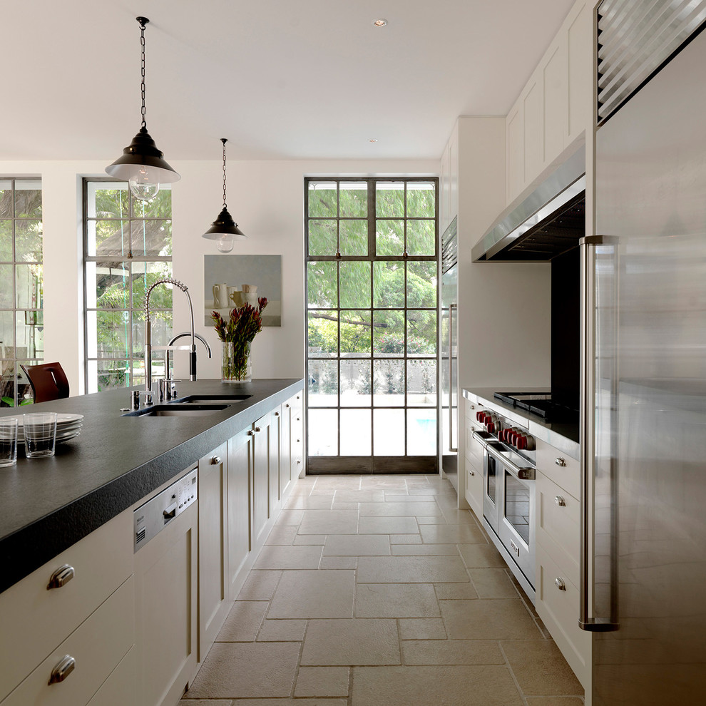 Cette image montre une cuisine parallèle traditionnelle avec un évier 2 bacs, un placard à porte shaker, des portes de placard blanches, un électroménager en acier inoxydable et un sol en travertin.