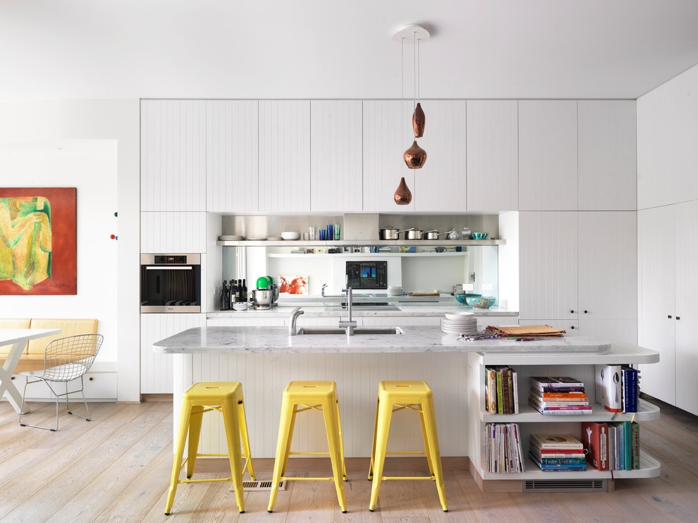 Bild på ett mellanstort funkis kök, med en undermonterad diskho, vita skåp, marmorbänkskiva, spegel som stänkskydd, rostfria vitvaror, ljust trägolv och en köksö