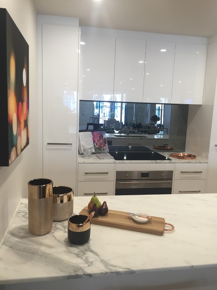 Bild på ett litet funkis kök, med en nedsänkt diskho, släta luckor, vita skåp, marmorbänkskiva, svart stänkskydd, spegel som stänkskydd, rostfria vitvaror, mellanmörkt trägolv och en köksö