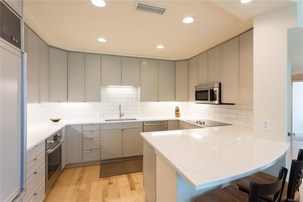 This is an example of a medium sized modern u-shaped kitchen/diner in Denver with a built-in sink, flat-panel cabinets, grey cabinets, composite countertops, white splashback, metro tiled splashback, stainless steel appliances, light hardwood flooring, a breakfast bar and brown floors.