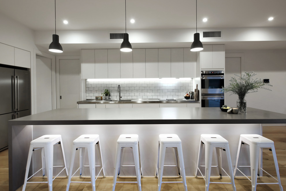 Photo of a contemporary l-shaped kitchen in Melbourne with an integrated sink, flat-panel cabinets, white cabinets, white splashback, stainless steel appliances, light hardwood flooring, an island, beige floors and black worktops.