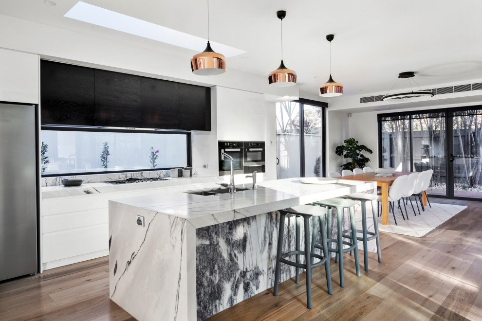 Photo of a large contemporary galley kitchen/diner in Melbourne with a double-bowl sink, flat-panel cabinets, white cabinets, marble worktops, glass sheet splashback, stainless steel appliances, medium hardwood flooring, an island, brown floors and grey worktops.